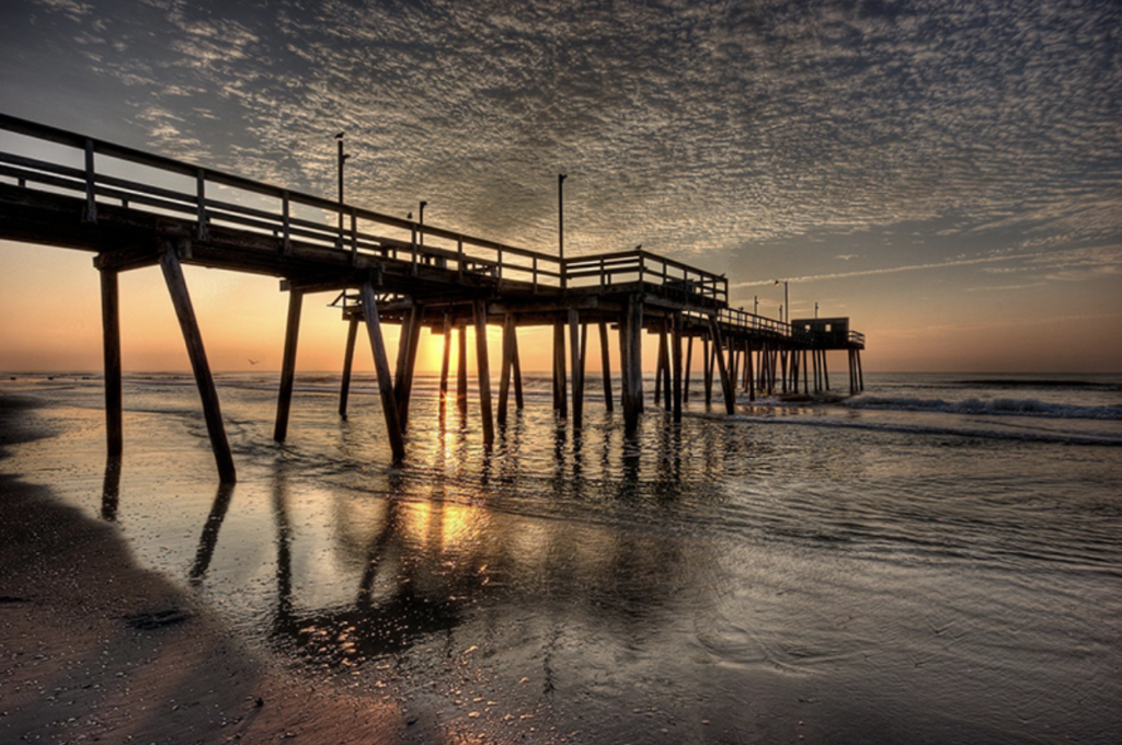 As the sun peeks over the horizon, the fishing pier stands silhouetted against the morning sky, its weathered planks basking in the golden hue.
